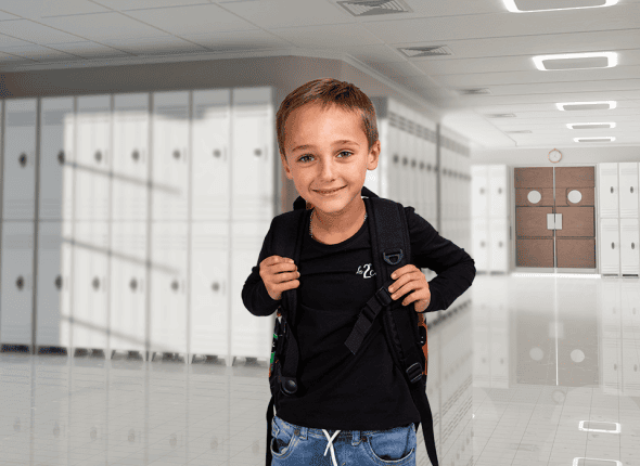 Un petit garçon avec un sac à dos se tient dans un couloir de l'école, prêt à commencer sa journée d'apprentissage. Son expression vive et curieuse montre son excitation face aux nouvelles découvertes qui l'attendent chez La 2e Classe.