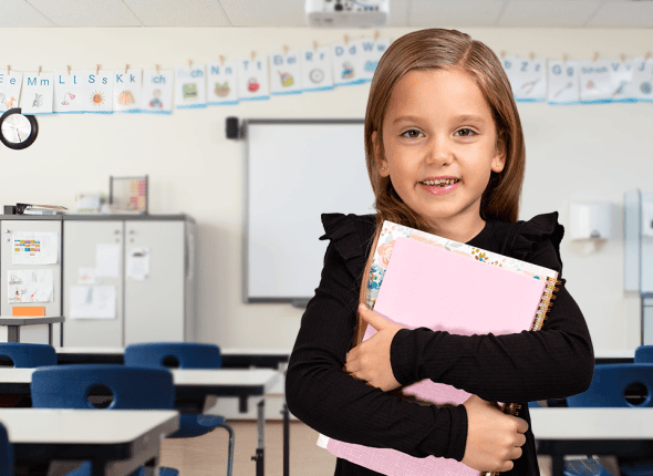 Une petite élève tient avec soin une pile de livres dans une salle de classe de La 2e Classe. Son expression joyeuse et curieuse reflète son enthousiasme pour l'apprentissage et la découverte de nouveaux savoirs.