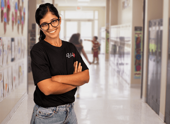 Une enseignante souriante se tient dans un couloir d'école lumineux, reflétant l'atmosphère accueillante et positive de La 2e Classe. Son sourire chaleureux montre sa passion pour l'éducation et son engagement envers le succès de chaque élève.