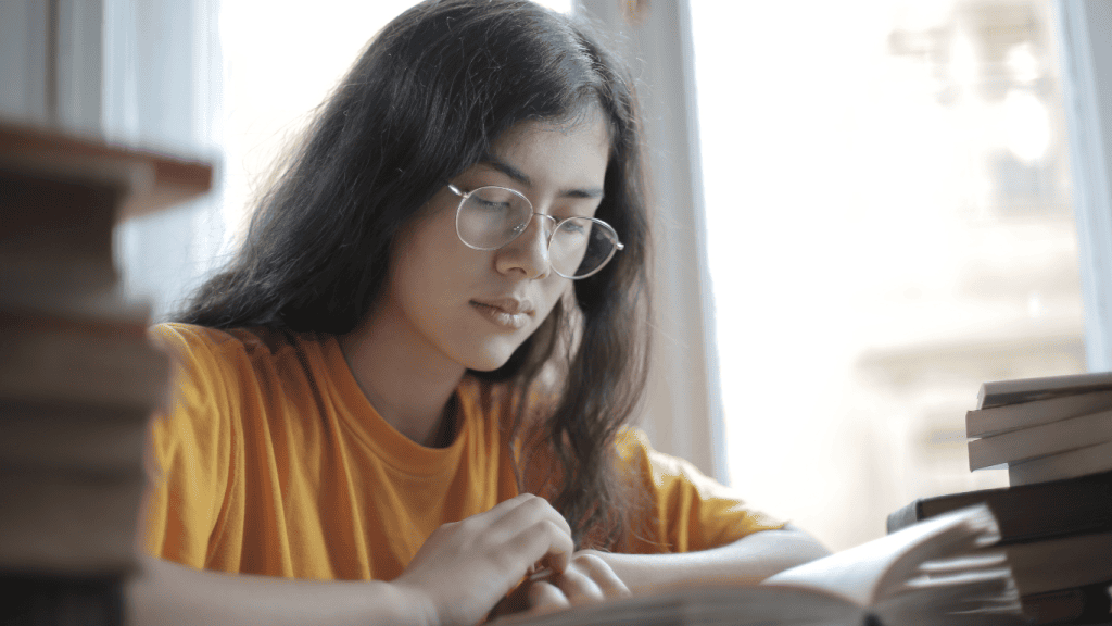 Jeune femme aux cheveux longs et lunettes, concentrée sur ses études, lisant un livre près d'une fenêtre, entourée de piles de livres. Étudiante en pleine révision dans un cadre lumineux.