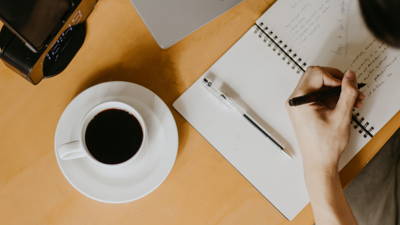 Main écrivant dans un cahier à côté d’une tasse de café et d’un stylo, sur un bureau.