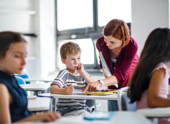 Enseignante accompagnant un jeune élève en classe pour l’aider à comprendre un exercice scolaire pendant que d’autres enfants travaillent.