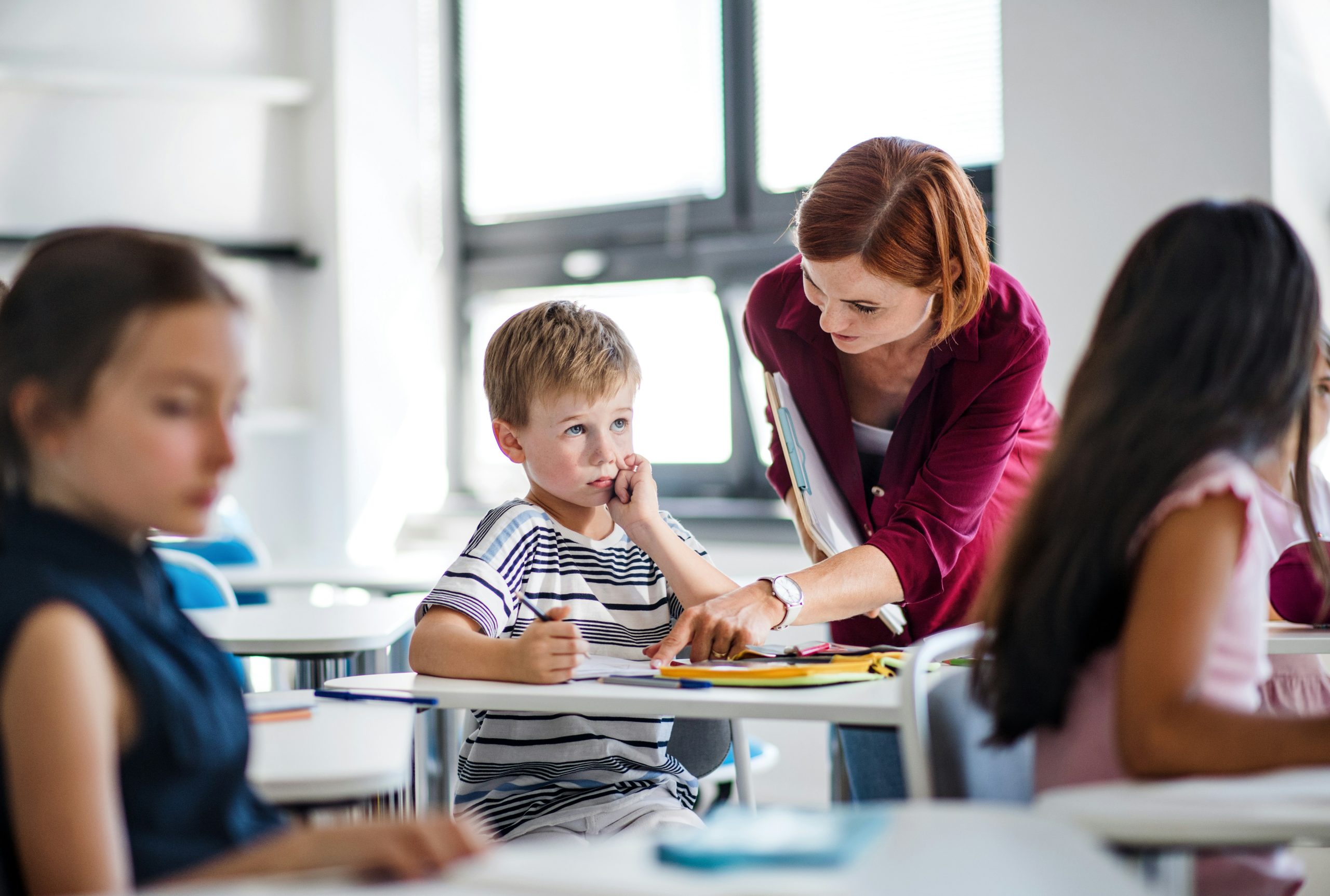 Enseignante accompagnant un jeune élève en classe pour l’aider à comprendre un exercice scolaire pendant que d’autres enfants travaillent.